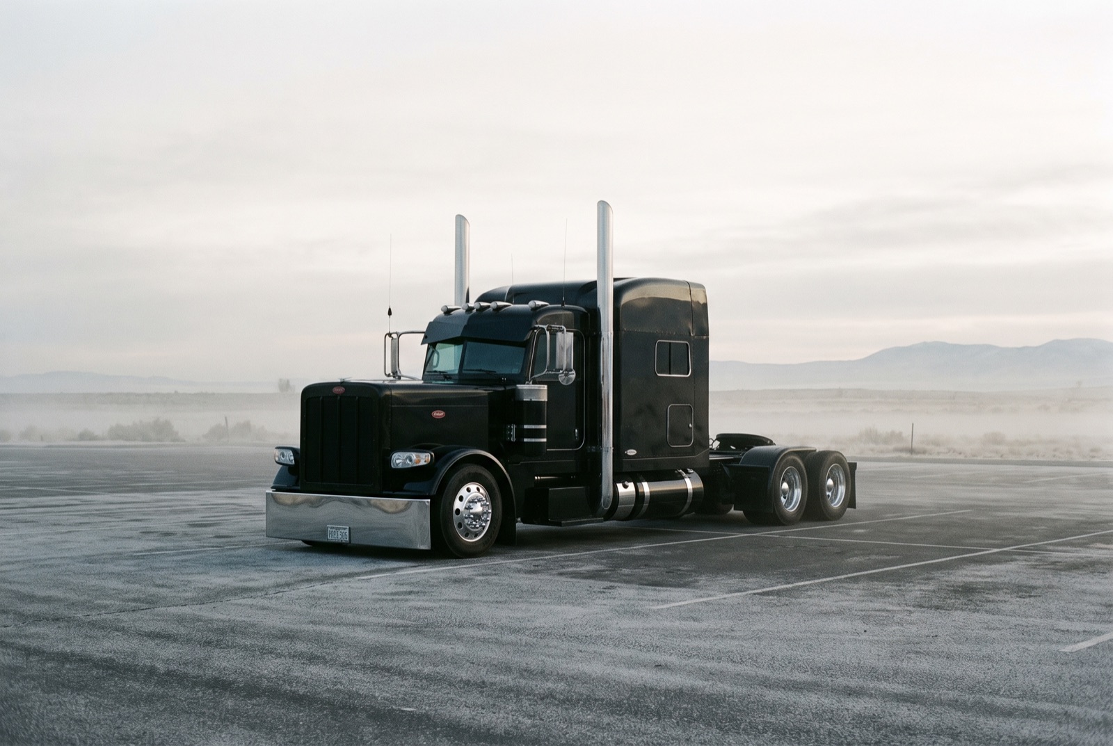 A blacked-out custom Peterbilt parked alone in an empty lot at cold misty dawn